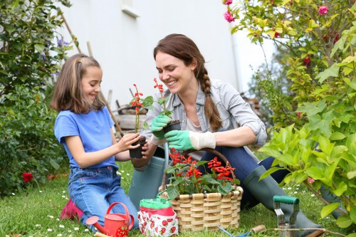 A woman and a young girl are gardening together in a lush outdoor space, likely in a residential garden near Harlesden. The woman, dressed in a light grey jacket and green gardening gloves, is kneeling on the grass and smiling as she helps the girl, who is wearing a blue t-shirt and pink shoes, plant a small flowering plant in a black pot. The garden background features dense green foliage, including various shrubs and flowering plants, with a white wall or fence partially visible. Nearby, there is a woven basket filled with gardening tools and a small red watering can, indicating active outdoor maintenance. The scene is set in natural daylight, possibly during a mild weather day, emphasizing a neat, well-maintained garden with a mix of grass, flower beds, and potted plants. This peaceful moment demonstrates typical gardening activities that Gardening Services Harlesden might provide, highlighting care for outdoor spaces within the local area, with vibrant natural tones of green, red, and earthy browns.