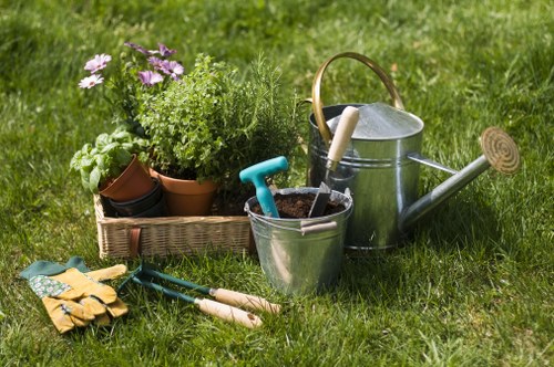 A well-maintained garden scene featuring a lush, green lawn with various gardening tools and equipment arranged in the foreground. There are multiple potted plants, including leafy greens and flowering plants, placed in a wicker basket, enhancing the natural and vibrant appearance. A metal watering can with a wooden handle and a small garden trowel are positioned nearby, ready for outdoor maintenance. Gardening gloves and pruning shears lie on the grass, indicating recent or upcoming gardening activities. The background showcases an evenly cut lawn extending toward a neatly edged flower bed or shrub border, contributing to a tidy and landscaped outdoor space. The lighting suggests a partly cloudy day with soft natural light illuminating the garden scene. This setting depicts a typical backyard garden prepared for ongoing plant care, with no visible structures or additional features, aligning with professional gardening and landscaping services offered in the Harlesden area, near London postcode NW10.