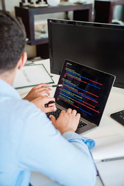 This image shows a person working at a desk with a laptop displaying lines of colorful code. The individual is holding a pen in one hand and appears to be engaged in programming or troubleshooting dig