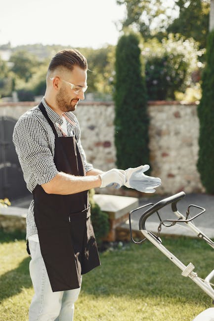 A man with styled dark hair, glasses, and a short beard is standing in a well-maintained garden, wearing a black apron over a checkered shirt and white trousers. He is putting on white gloves in prepa