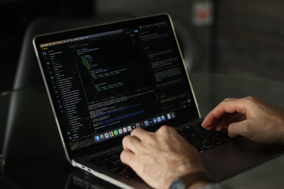 A close-up view of a person's hands operating a laptop with a dark screen displaying lines of colorful code, situated on a reflective black surface. The background is blurred, suggesting an indoor env